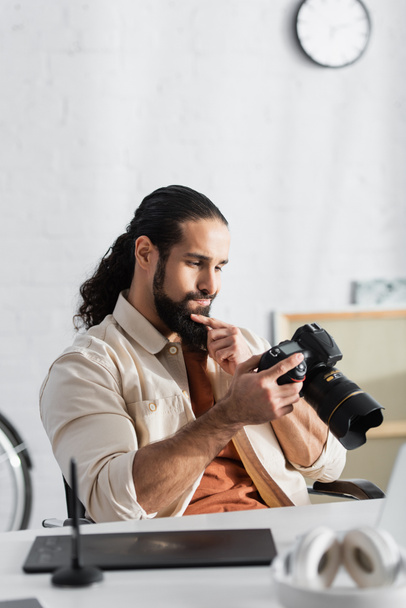 przemyślany Hiszpan dotykający brody patrząc na aparat cyfrowy w pobliżu rozmytej tablicy graficznej - Zdjęcie, obraz