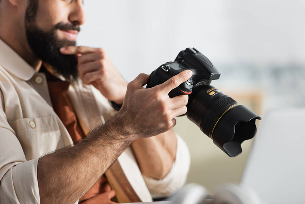 обрезанный вид вдумчивого человека, трогающего бороду, держа цифровую камеру дома - Фото, изображение