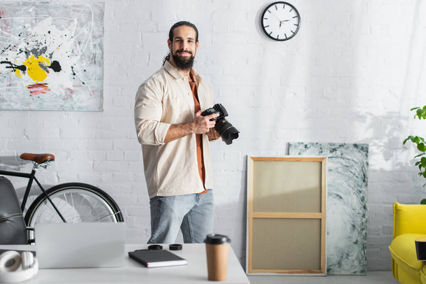 hispánec s digitálním fotoaparátem stojící v moderním obývacím pokoji v blízkosti stolu s rozmazaným notebookem - Fotografie, Obrázek