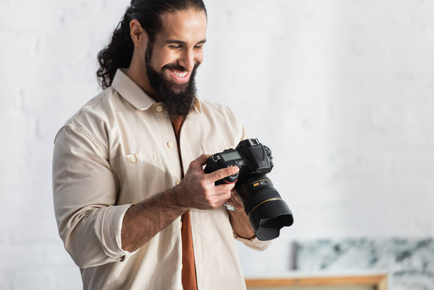 heureux homme hispanique souriant tout en regardant appareil photo numérique moderne à la maison - Photo, image