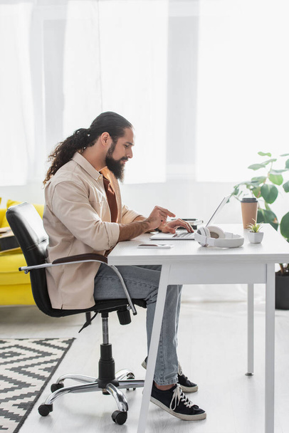 vista lateral de barbudo latino freelancer digitando no laptop enquanto trabalhava em casa - Foto, Imagem