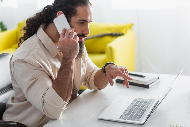 szakállas spanyol szabadúszó laptopra mutat, miközben otthon okostelefonon beszél. - Fotó, kép