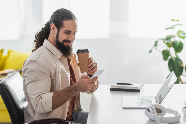 smiling hispanic freelancer with coffee to go messaging on smartphone near laptop at home - Photo, Image