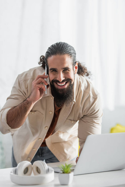 alegre freelancer hispano mirando a la cámara mientras habla en el teléfono celular cerca de la computadora portátil borrosa - Foto, imagen