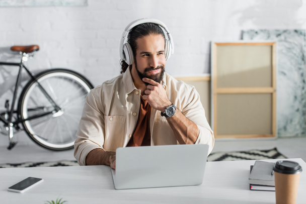 χαμογελαστός ισπανόφωνος ελεύθερος επαγγελματίας με ακουστικά που σκέφτεται κοντά στο φορητό υπολογιστή στο σπίτι - Φωτογραφία, εικόνα