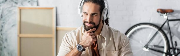 uśmiechnięty i troskliwy Hiszpan dotykający brody podczas słuchania muzyki w słuchawkach, sztandar - Zdjęcie, obraz
