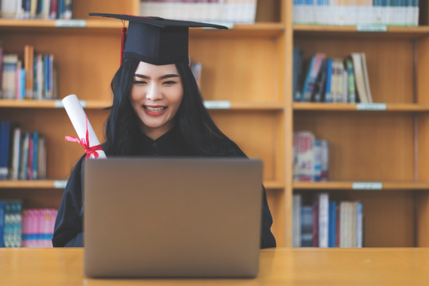 University graduate in graduation gown and mortarboard celebrates in a virtual graduation ceremony. Happy female student on her graduation day at home. Concept of online education - Photo, Image