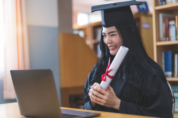 University graduate in graduation gown and mortarboard celebrates in a virtual graduation ceremony. Happy female student on her graduation day at home. Concept of online education - Photo, Image