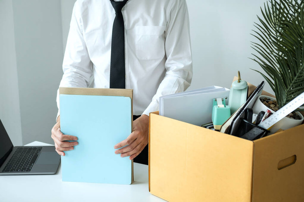 Работник компании упаковывает документы и забирает личные вещи в коричневую картонную коробку, чтобы покинуть рабочее место при увольнении с работы или смене работы - Фото, изображение