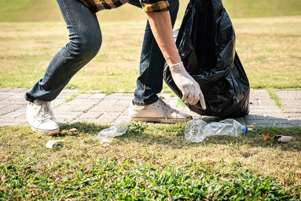Vrijwilliger in handschoenen lopen en stoppen om plastic flessen te verzamelen in plastic zwarte zak voor het reinigen van het park tijdens de milieuactiviteit tot het verzamelen van afval - Foto, afbeelding