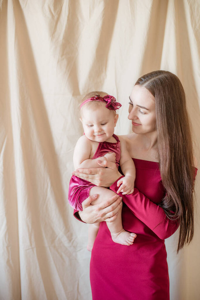 A young mother with long dark hair in a burgundy dress with a baby in her arms on a light background. Healthy lifestyle. Happy motherhood  - Photo, Image