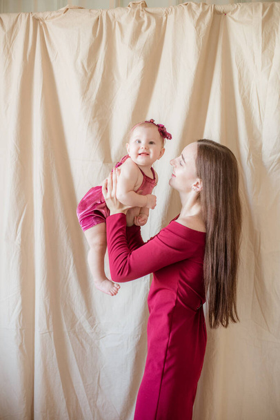 A young mother with long dark hair in a burgundy dress with a baby in her arms on a light background. Healthy lifestyle. Happy motherhood  - Photo, image