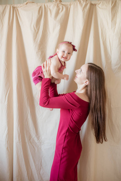 Une jeune mère aux longs cheveux foncés en robe bordeaux avec un bébé dans les bras sur un fond clair. Mode de vie sain. Bonne maternité  - Photo, image