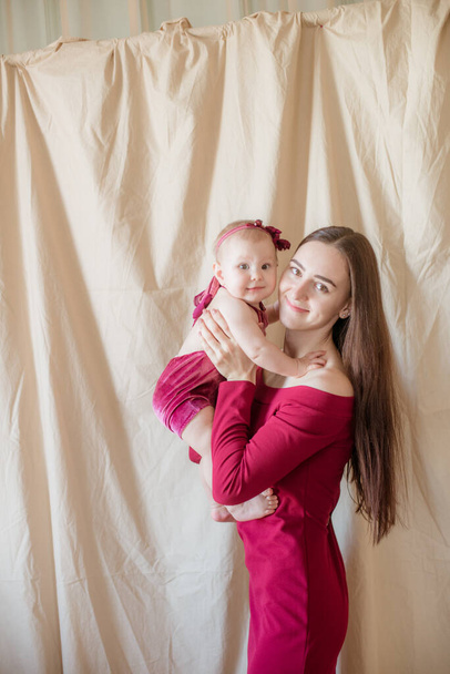 A young mother with long dark hair in a burgundy dress with a baby in her arms on a light background. Healthy lifestyle. Happy motherhood  - Foto, Bild