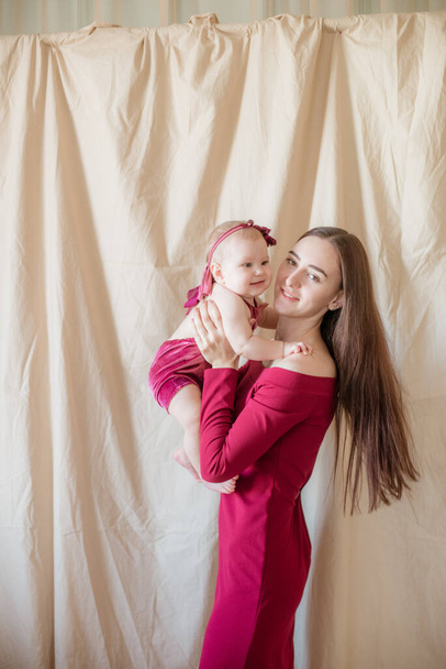 A young mother with long dark hair in a burgundy dress with a baby in her arms on a light background. Healthy lifestyle. Happy motherhood  - Photo, image