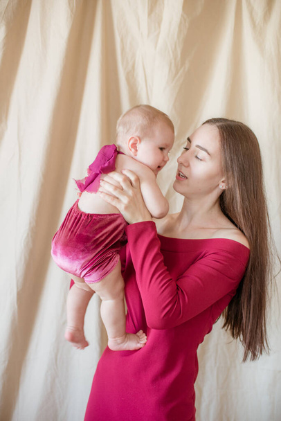 A young mother with long dark hair in a burgundy dress with a baby in her arms on a light background. Healthy lifestyle. Happy motherhood  - Fotó, kép
