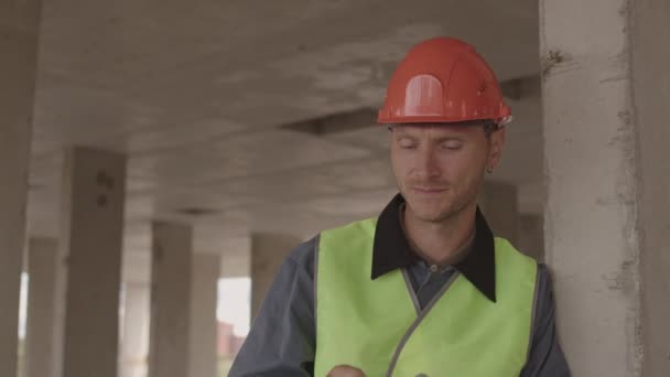 Грудь белого мужчины средних лет в рефлекторном жилете, снимает и надевает каску уставшей после тяжелой работы, стоит в строящемся здании, опираясь на бетонную колонну - Кадры, видео