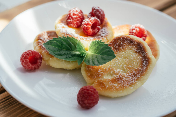 Delicious cheesecakes or pancakes sprinkled with powdered sugar on white plate on wooden table with raspberries and mint leaf - Photo, Image