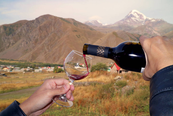 Hand pouring red wine into wineglass with blurry Mt. Kazbek in the backdrop, Goergia - Photo, Image