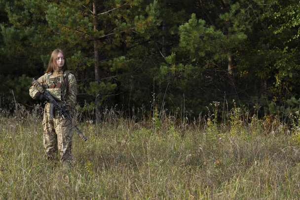 Uma menina com cabelos vermelhos em roupas de camuflagem e um colete à prova de balas fica em um campo contra o fundo de uma floresta. Uma mulher militar tem uma espingarda automática nas mãos. - Foto, Imagem
