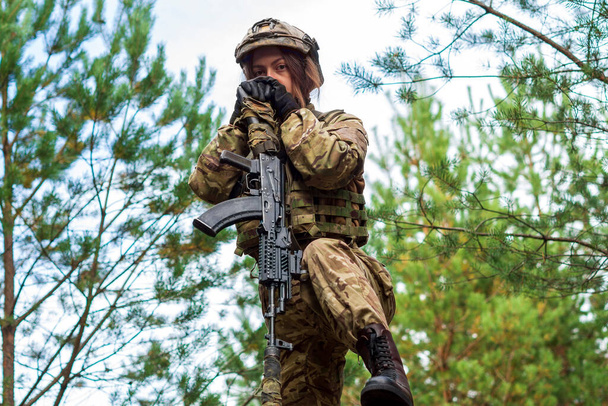 Retrato de uma jovem em um uniforme de camuflagem militar com uma espingarda automática em suas mãos contra o fundo de uma floresta. Uma mulher em um capacete tático e colete à prova de balas segura uma arma. - Foto, Imagem