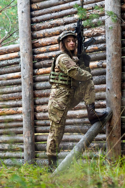 A rapariga de uniforme militar tem uma espingarda automática nas mãos. Mulher soldado em colete à prova de balas e capacete tático na floresta contra fundo de parede de madeira. Campo de treino, campo de tiro. - Foto, Imagem