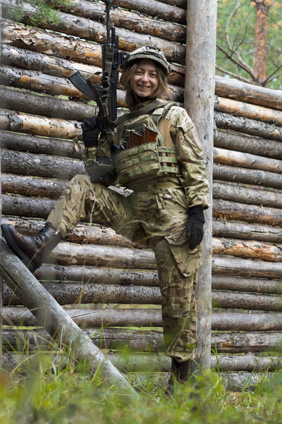 A rapariga de uniforme militar tem uma espingarda automática nas mãos. Mulher soldado em colete à prova de balas e capacete tático na floresta contra fundo de parede de madeira. Campo de treino, campo de tiro. - Foto, Imagem