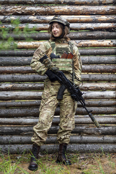 Egy katonai egyenruhás lány automata fegyvert tart a kezében. Női katona golyóálló mellényben és taktikai sisak az erdőben háttal fa fal. Edzőtábor, lőtér. - Fotó, kép