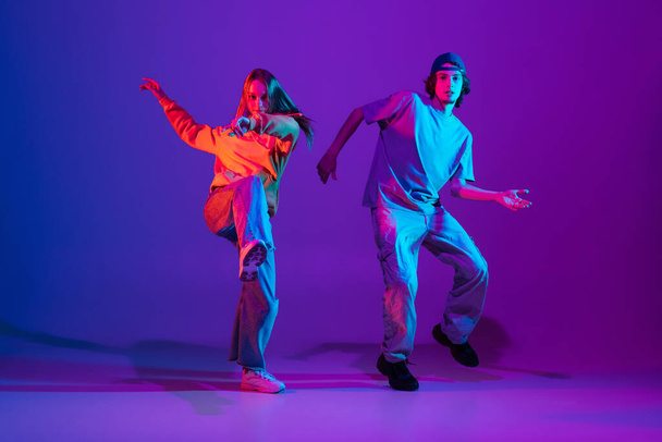 Two dancers, young man and woman dancing hip-hop in casual sports youth clothes on gradient purple pink background at dance hall in neon light. - Фото, зображення