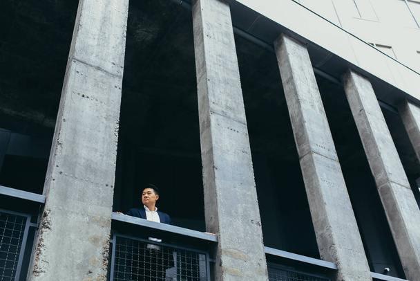 Genç Asyalı işadamı betondan yapılmış modern siyah ofis binasının yanında duruyor, balkon terasında sütunlarla birlikte. - Fotoğraf, Görsel