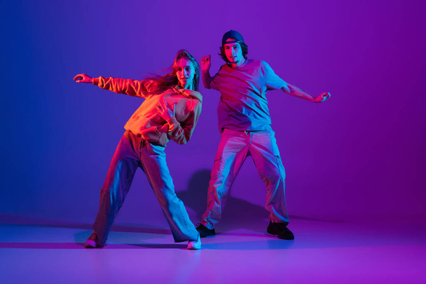 Young man and woman dancing hip-hop in casual sports youth clothes on gradient purple pink background at dance hall in neon light. - Fotoğraf, Görsel