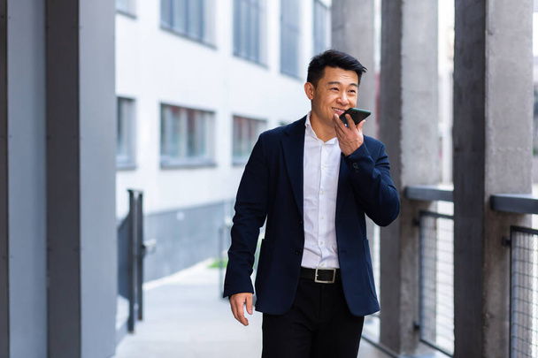 cheerful smiling Successful Asian businessman explains information to employees using phone, speaks near office outside happy talking - Foto, Imagen