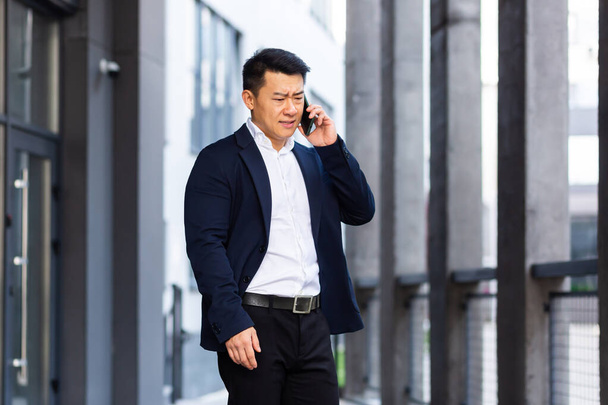 angry and serious Successful Asian businessman explains information to employees using phone, speaks near office outside - Photo, image