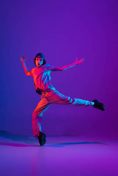 Young stylish man, break dancing, hip-hop dancer practicing solo in casual clothes isolated over purple background in neon pink light at dance hall - Foto, Imagen