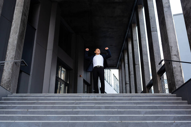Щасливий і веселий азіатський бізнесмен тримає руки вгору радіючи перемозі і успіху біля офісного центру вид спереду
 - Фото, зображення