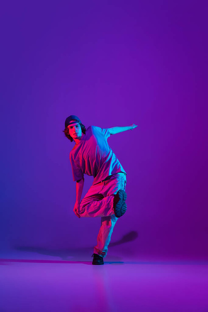 Stylish sportive boy dancing hip-hop in stylish clothes on colorful background at dance hall in neon light. Youth culture, movement, style and fashion, action. - Photo, image