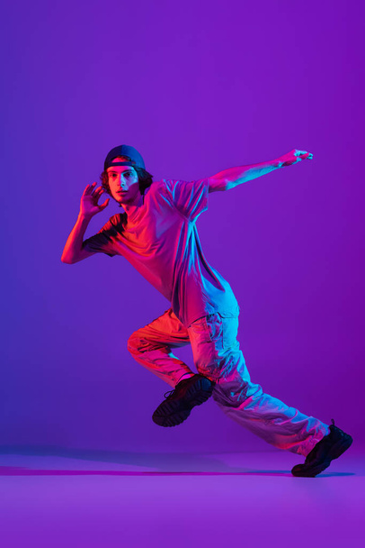 Young stylish man, break dancing, hip-hop dancer practicing solo in casual clothes isolated over purple background in neon pink light at dance hall - 写真・画像