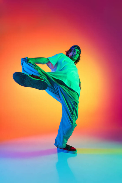 Conceptual portrait of young man, hip-hop dancer in stylish clothes in action isolated on colorful background at dance hall in neon light. - 写真・画像