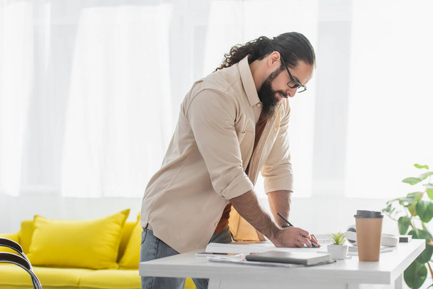 diseñador hispano sosteniendo lápiz mientras está de pie cerca del escritorio en casa - Foto, Imagen