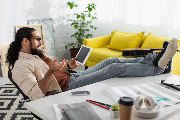 designer latino surpris assis avec les jambes sur le bureau et geste près de tablette numérique - Photo, image