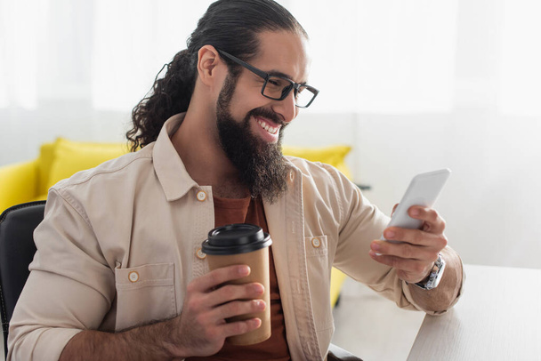 Χαρούμενος ισπανόφωνος άνδρας με γυαλιά ηλίου κρατώντας καφέ για να πάει και να χρησιμοποιήσει smartphone στο σπίτι - Φωτογραφία, εικόνα
