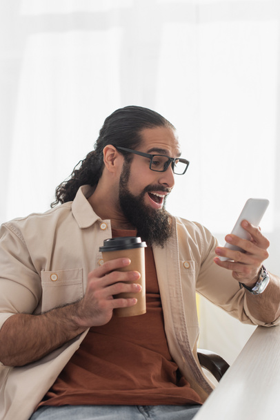 asombrado hombre hispano con café para ir mirando el teléfono inteligente en casa - Foto, imagen
