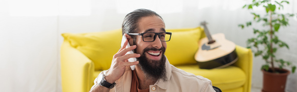 hombre hispano feliz hablando en el teléfono inteligente cerca del sofá y la guitarra acústica sobre fondo borroso, bandera - Foto, imagen