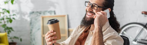 usmívající se latinskoamerický muž v brýlích, který drží kávu s sebou, zatímco doma mluví na mobilu, banner - Fotografie, Obrázek