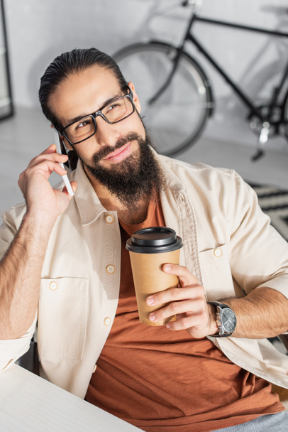 hombre latino sonriendo mientras sostiene la bebida para llevar y llamando al teléfono celular cerca de la bicicleta borrosa en casa - Foto, Imagen