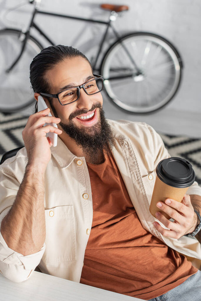 gioioso uomo latino con caffè per andare a parlare su smartphone vicino alla bici su sfondo sfocato a casa - Foto, immagini