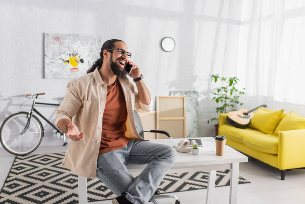 laughing hispanic man talking on smartphone while sitting on desk in modern living room - Photo, Image