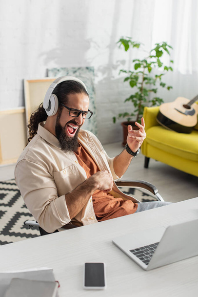 emocionado freelancer hispano en auriculares gritando mientras escucha música cerca de la computadora portátil - Foto, Imagen