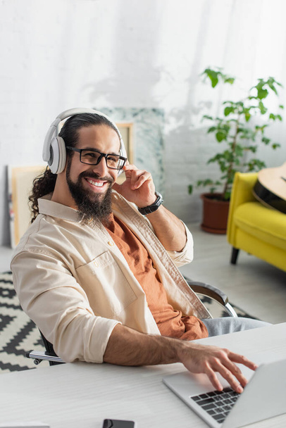 bearded hispanic freelancer in headphones smiling at camera while listening music near blurred laptop - Fotó, kép