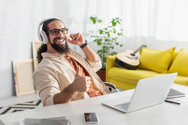 vrolijk latijn man tonen duim omhoog tijdens het luisteren van muziek in hoofdtelefoons in de buurt van laptop op het bureau - Foto, afbeelding